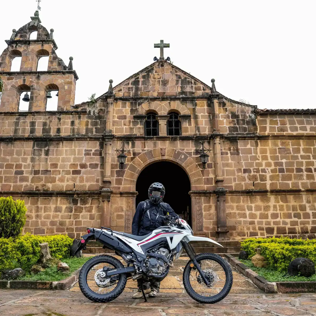 Rider on a motorcycle in front of a stone church with a bell tower and an arched doorway