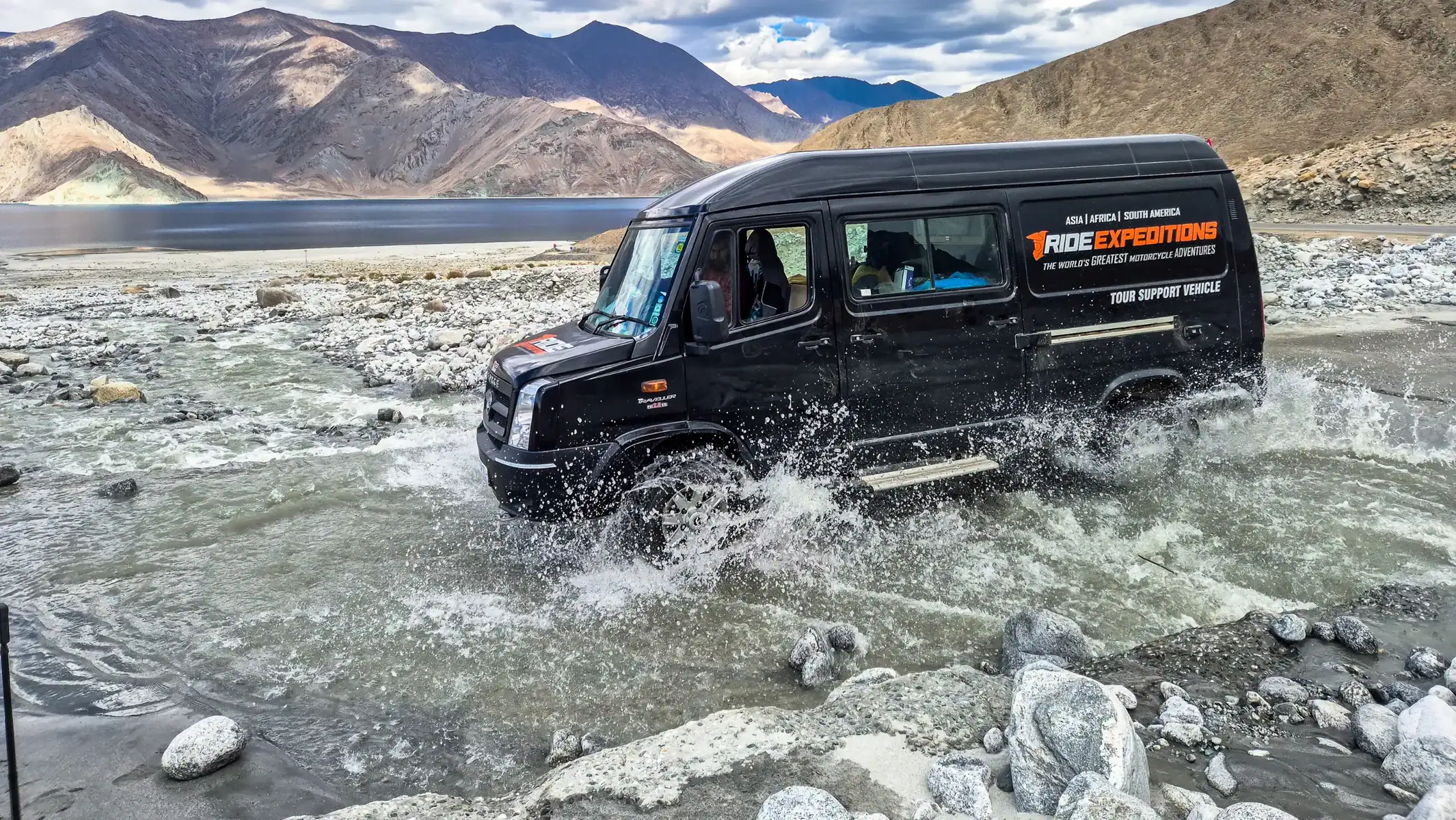 Black minibus with Ride Expeditions lettering crossing a shallow rocky stream